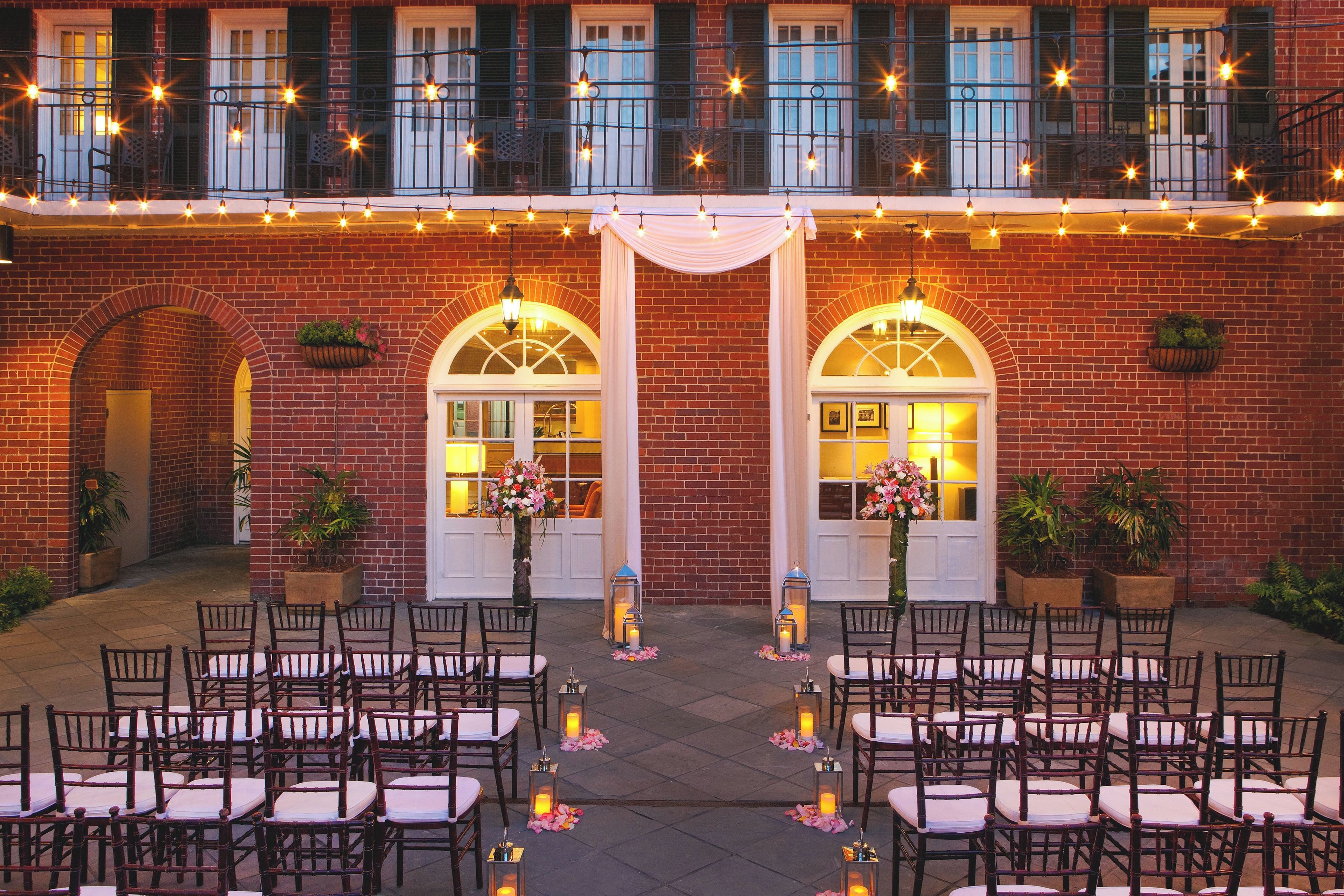 An outdoor seating area with chairs and tables, a brick building with arched doorways, and string lights above.