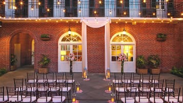 An outdoor seating area with chairs and tables, a brick building with arched doorways, and string lights above.
