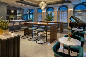 A spacious bar area with high ceilings, a grand piano, and multiple seating options including bar stools and round tables.