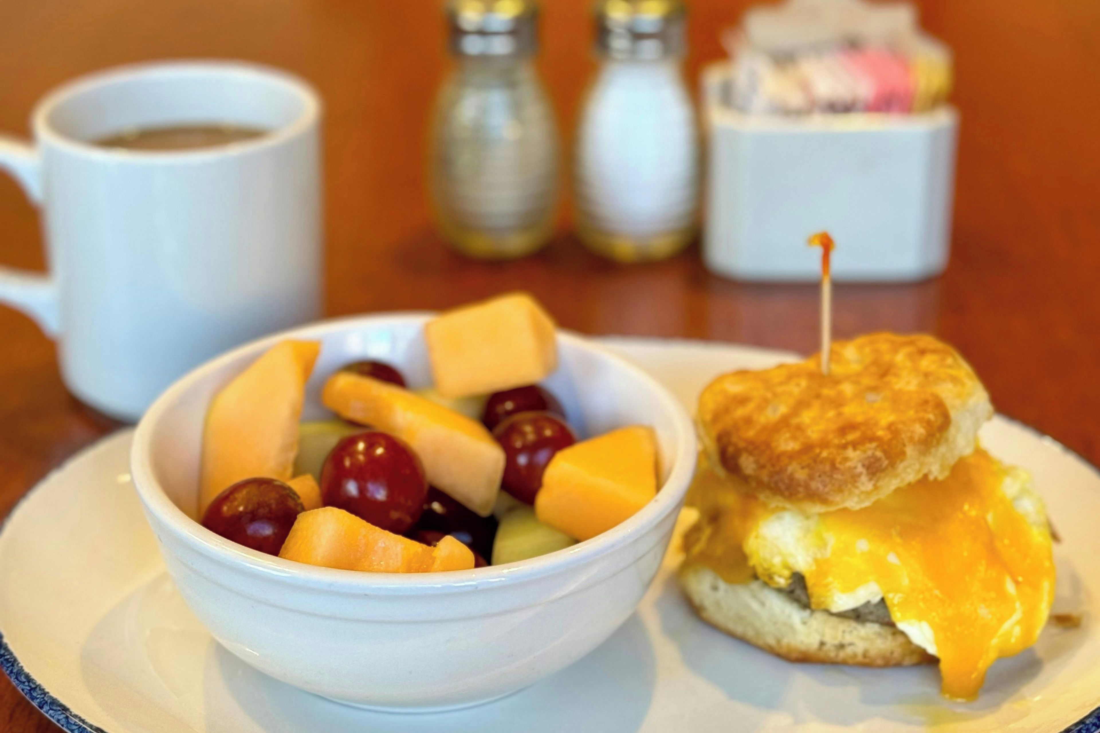 A breakfast plate with a bowl of fruit, a sandwich, and a cup of coffee.