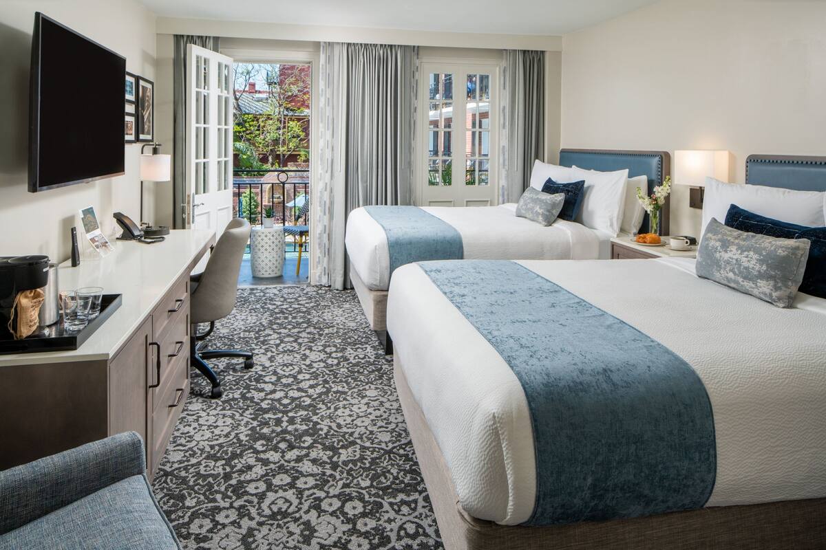 Quarto de hotel com duas camas, uma escrivaninha, uma cadeira, televisão e varanda com cortinas.