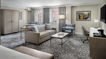 Quarto de hotel com sofá, poltrona, escrivaninha e uma janela grande com cortinas.