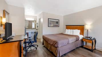 A hotel room with a bed, desk, chair, and a small table with a lamp.