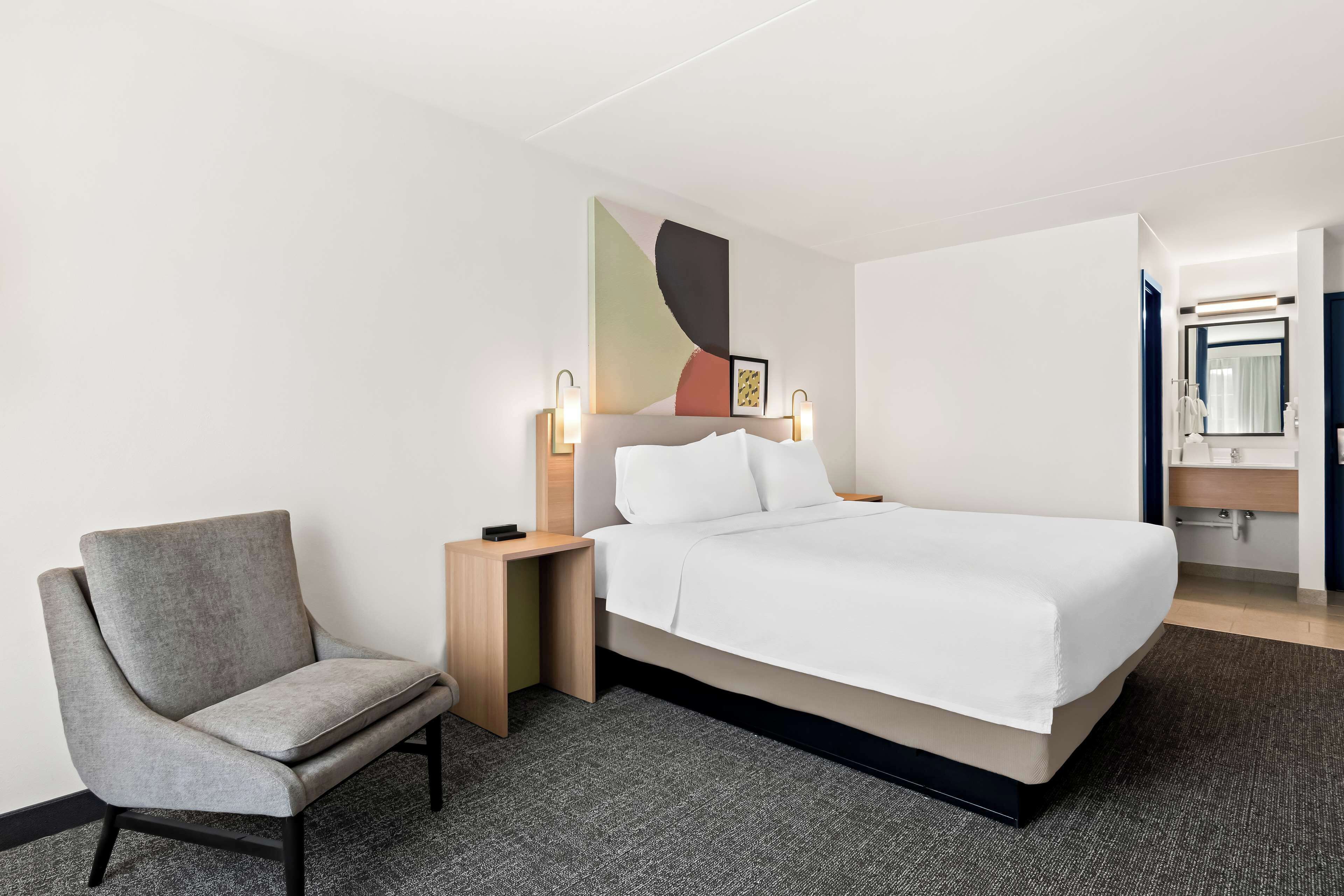 Une chambre d’hôtel avec un lit, une chaise, une table de chevet, une lampe fixée au mur et une salle de bain visible à travers une porte ouverte.