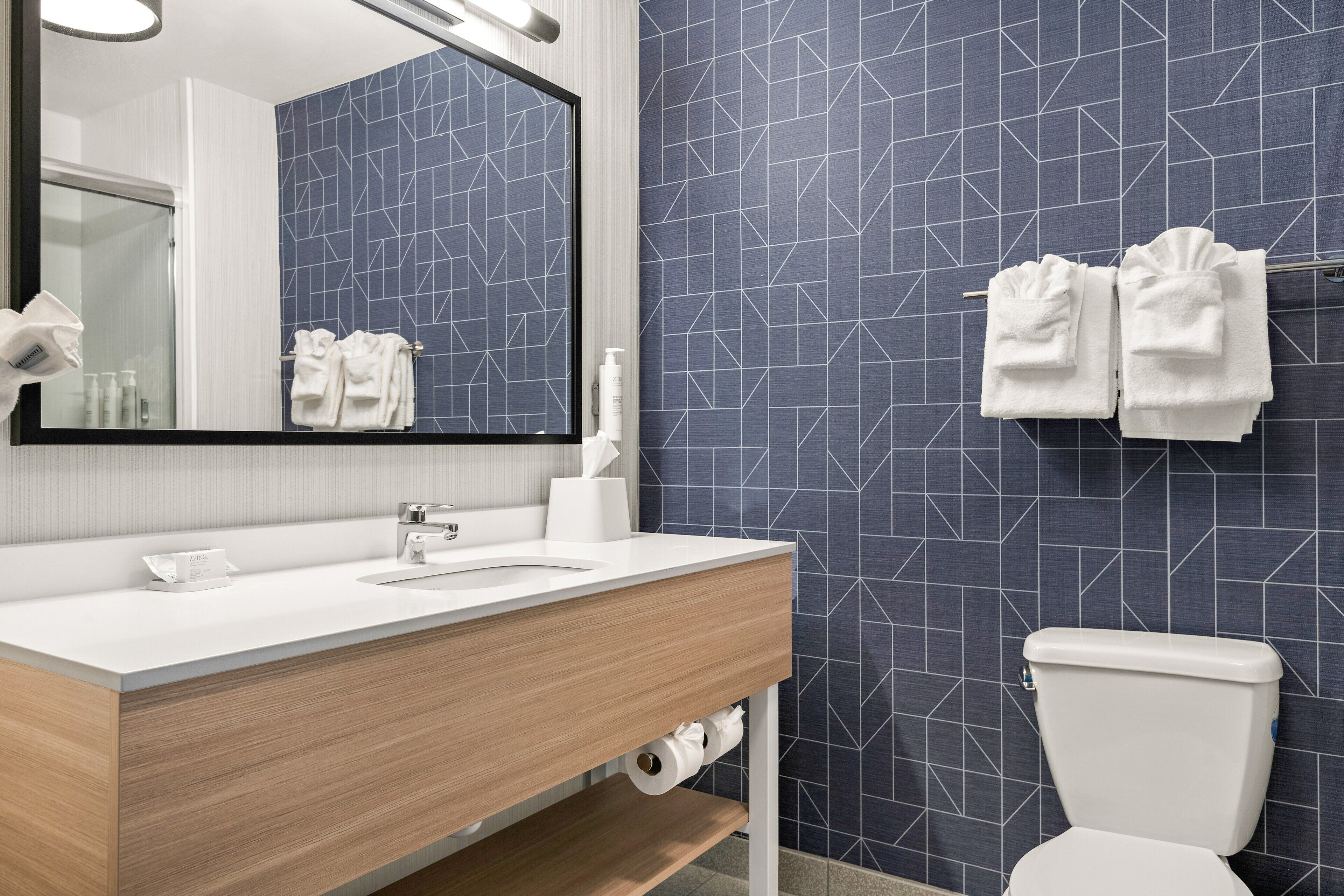 Een moderne badkamer met een witte wastafel, spiegel en toilet. Handdoeken hangen aan een rek en er is een toiletrolhouder aan de muur.