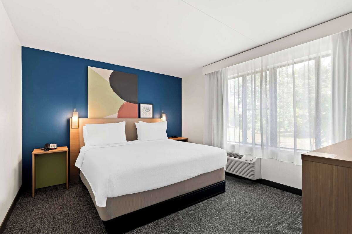 Une chambre d’hôtel avec un grand lit, un bureau, une table de chevet, une œuvre d’art murale et une fenêtre avec des rideaux légers.