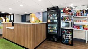 A reception area with a wooden counter, a refrigerator stocked with beverages, and shelves with snacks and condiments. - Spark by Hilton Hummelstown Hershey (Hummelstown)