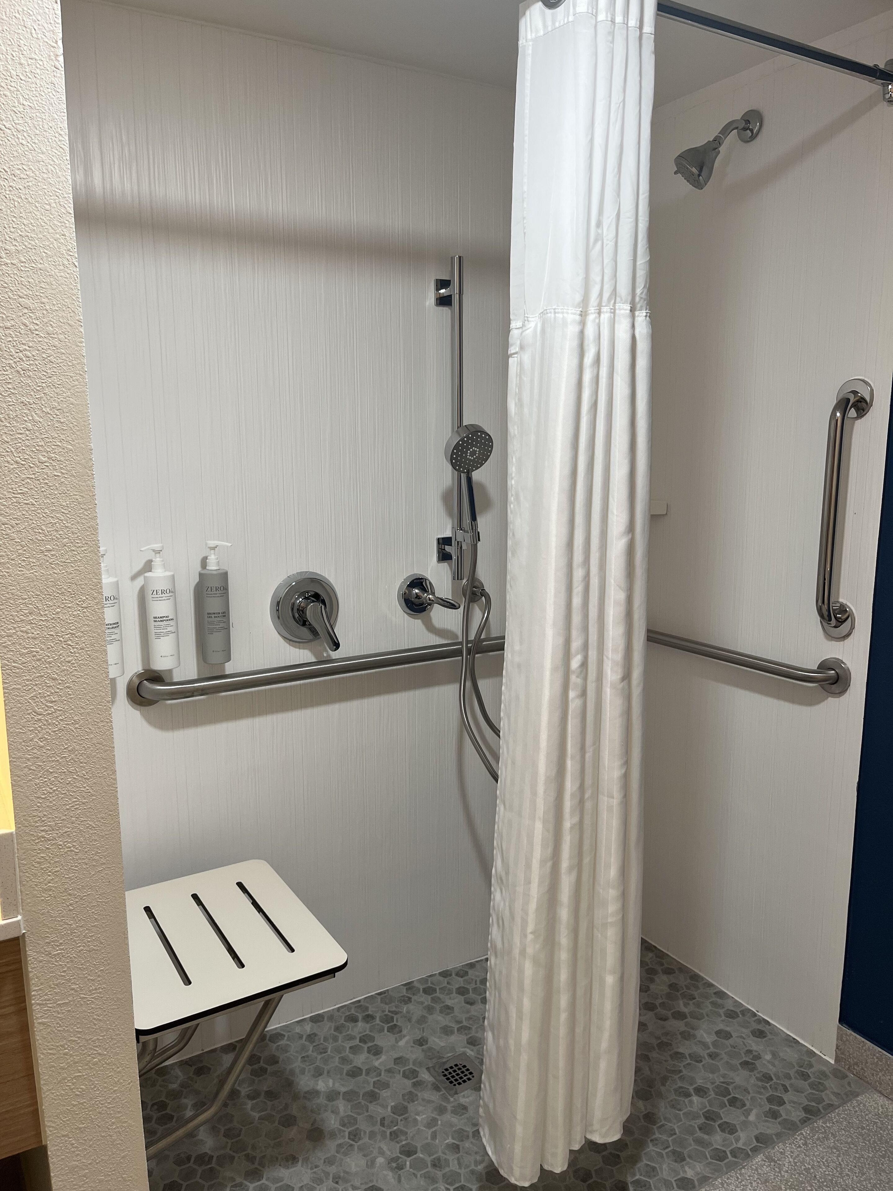 A shower area with a white shower curtain, grab bars, a handheld showerhead, and a handheld faucet.