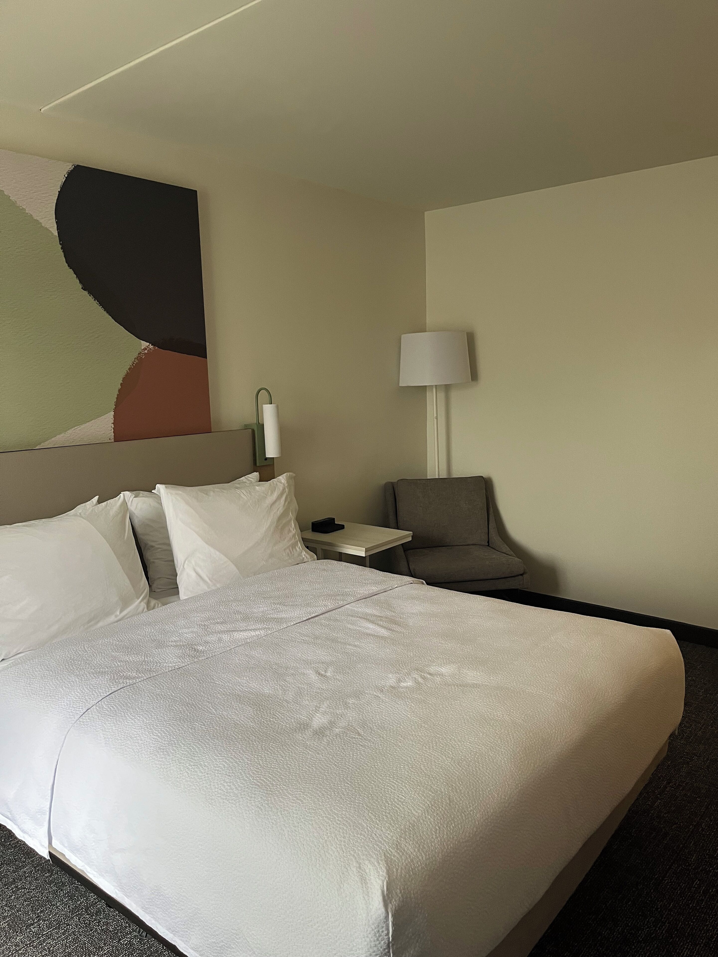 Une chambre d’hôtel avec un grand lit, une petite table, une chaise, une lampe et un mur orné d’un tableau aux motifs géométriques.