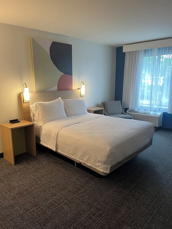 Una habitación de hotel con una cama grande, dos mesitas de noche, una silla, una ventana con cortinas y una obra de arte en la pared.