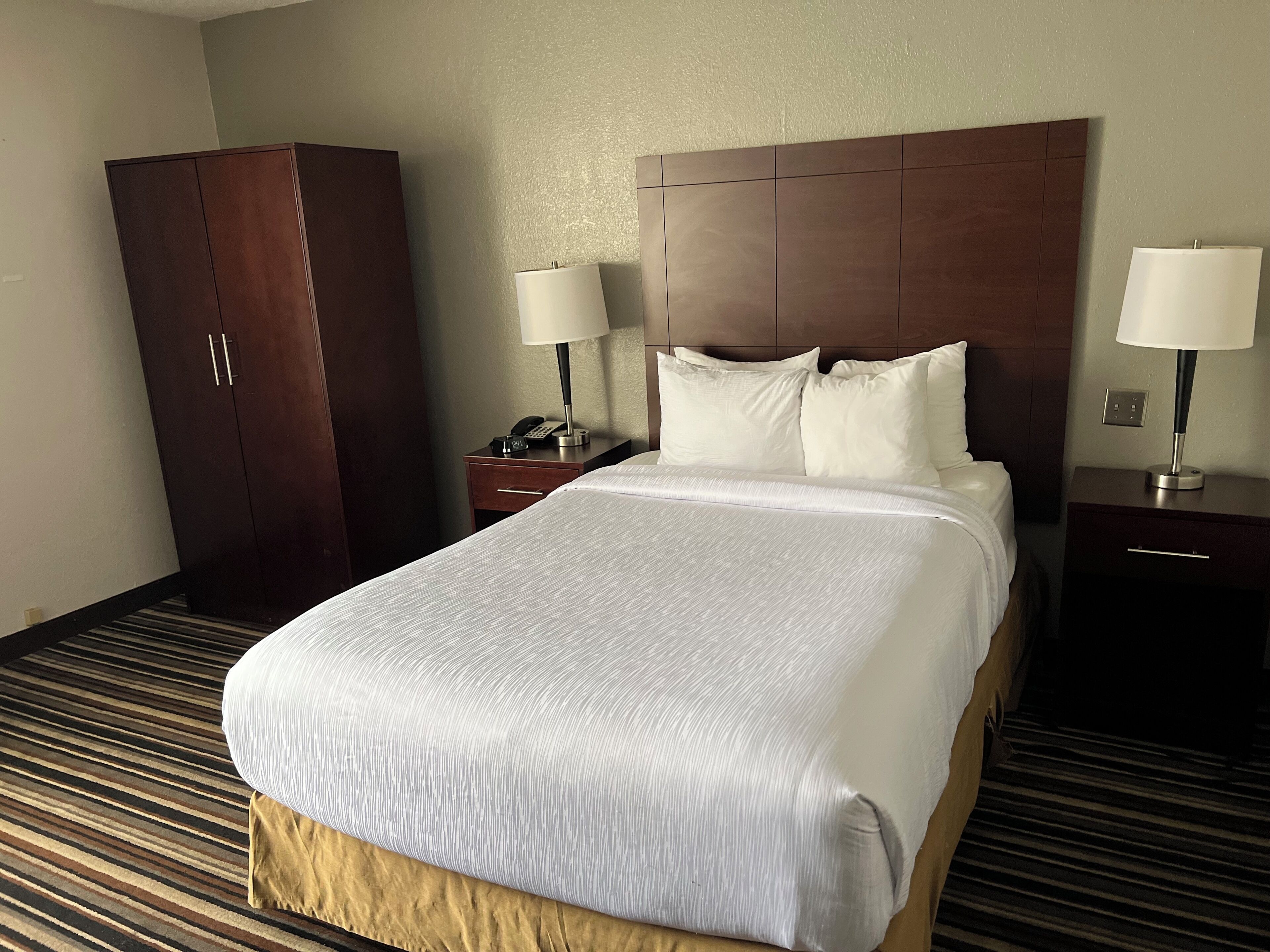 A hotel room with a large bed, two bedside tables with lamps, a wardrobe, and a carpeted floor.