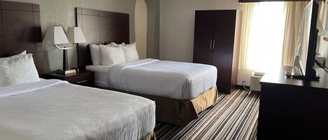 Quarto de hotel com duas camas, uma escrivaninha, luminária, armário e janela com cortinas.