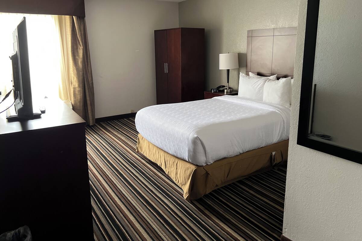 Une chambre d’hôtel avec un grand lit, une armoire, un bureau avec une télévision et une table de chevet avec une lampe.