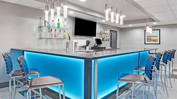 Un área de bar moderna con una barra iluminada en azul, varios taburetes, un televisor de pantalla plana y una variedad de botellas en estantes.