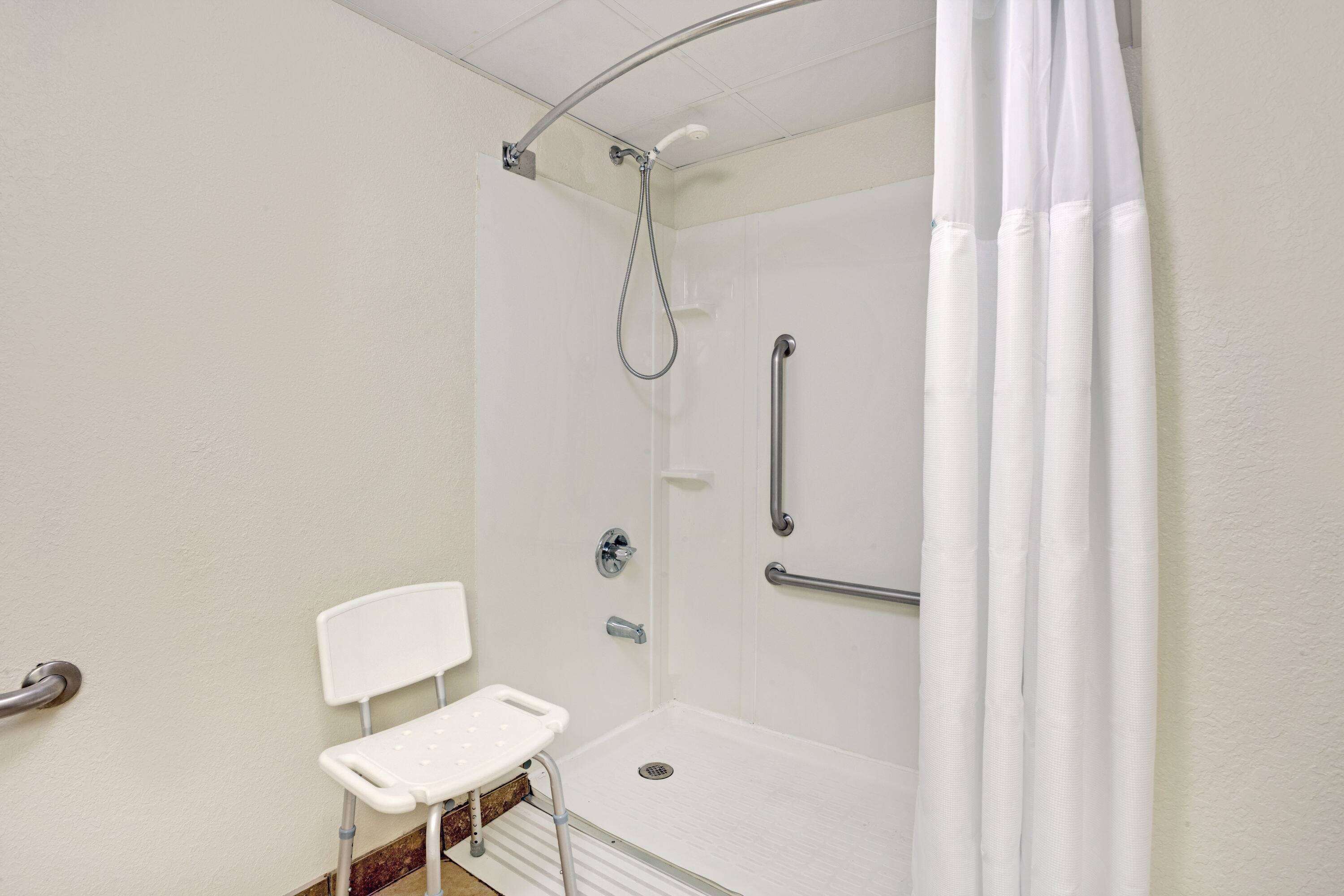 A white, accessible shower room with a grab bar, a white chair, and a showerhead.