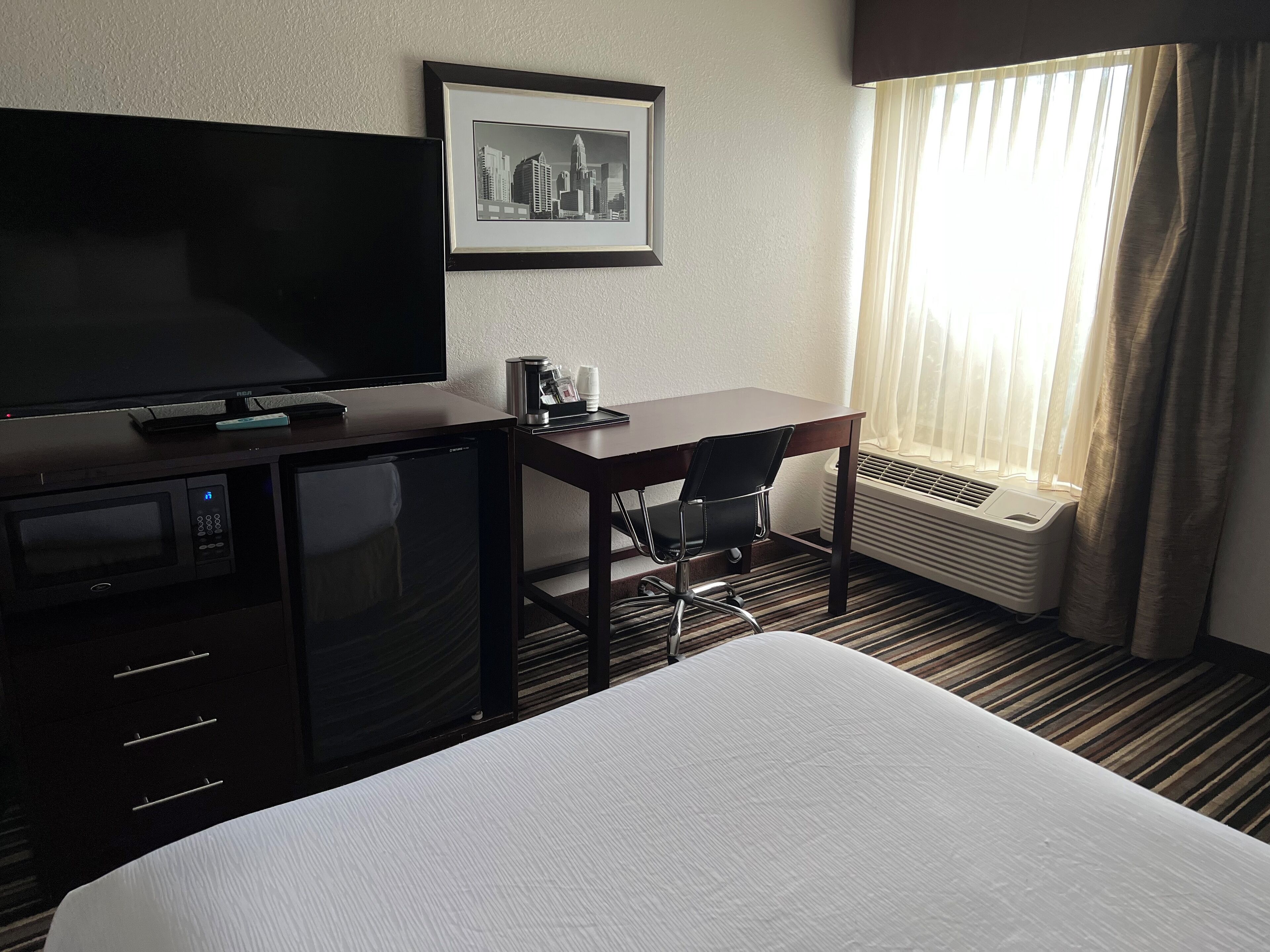 A hotel room with a bed, a desk with a chair, a television, a framed picture on the wall, and a window with curtains.