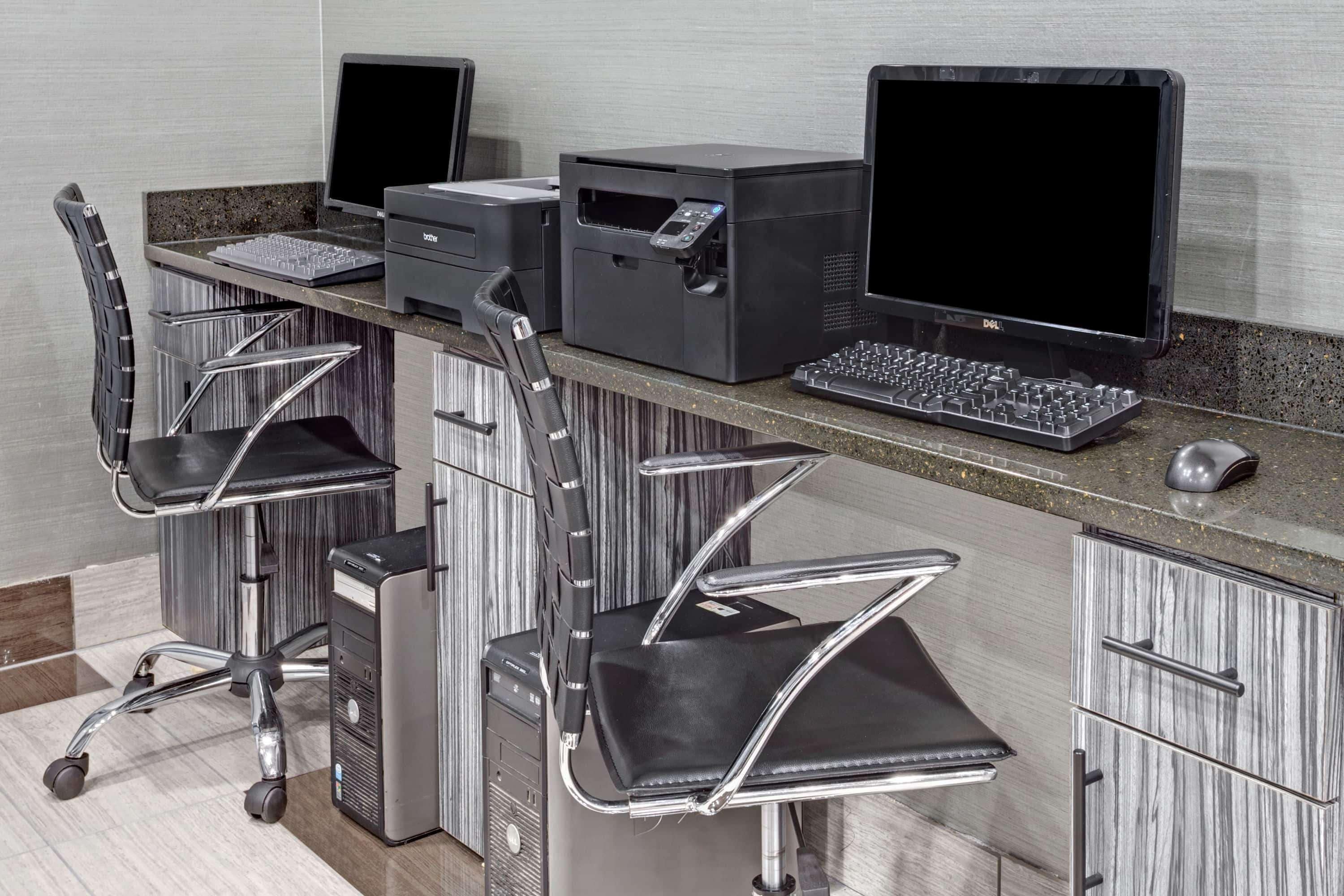 A modern office setup with two desks, each equipped with a computer monitor, keyboard, and printer. There are two ergonomic chairs and a small desk fan.