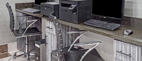 Un espacio de oficina moderno con dos escritorios, cada uno con monitor de computadora, teclado e impresora. Hay dos sillas ergonómicas y un pequeño ventilador de escritorio.