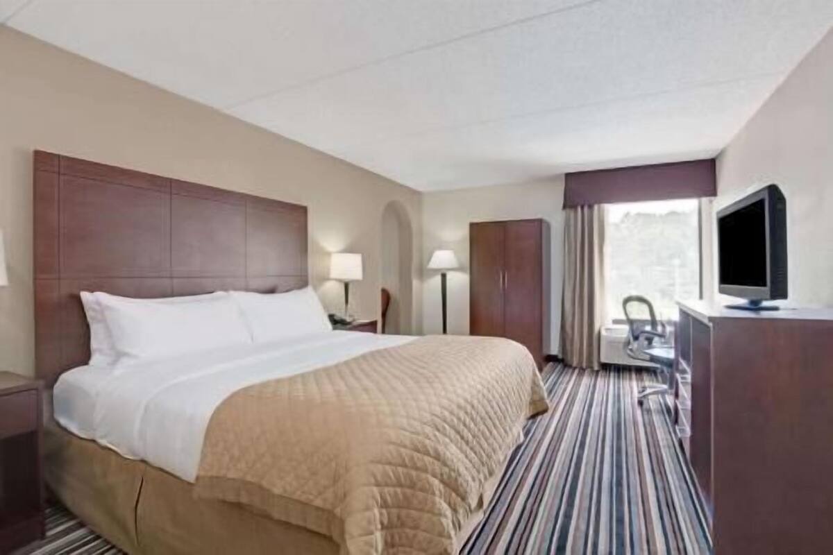 Une chambre d’hôtel avec un lit, un bureau avec un ordinateur, une télévision et une fenêtre avec des rideaux.