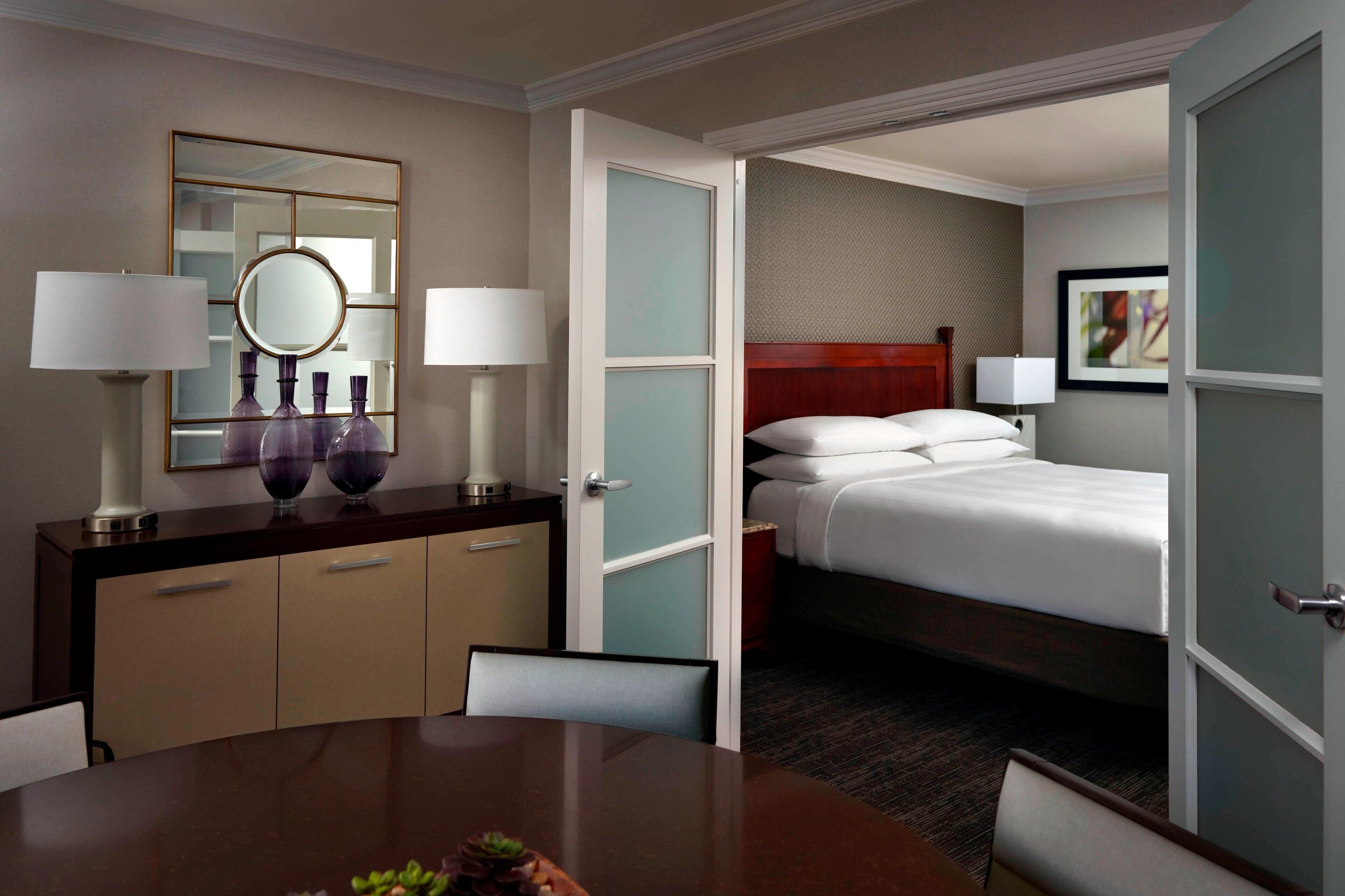 A hotel room with a bed, bedside table, lamp, mirror, and a view into another room.