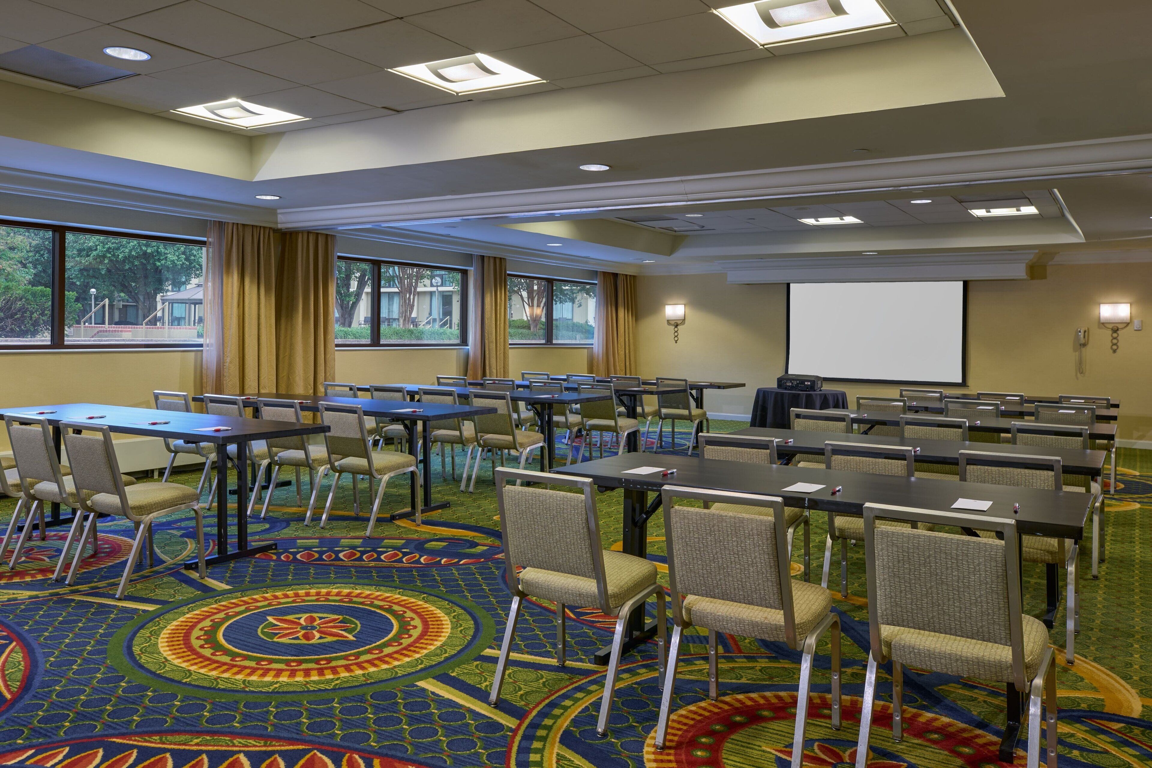 A conference room with tables and chairs, large windows, and a projector screen.