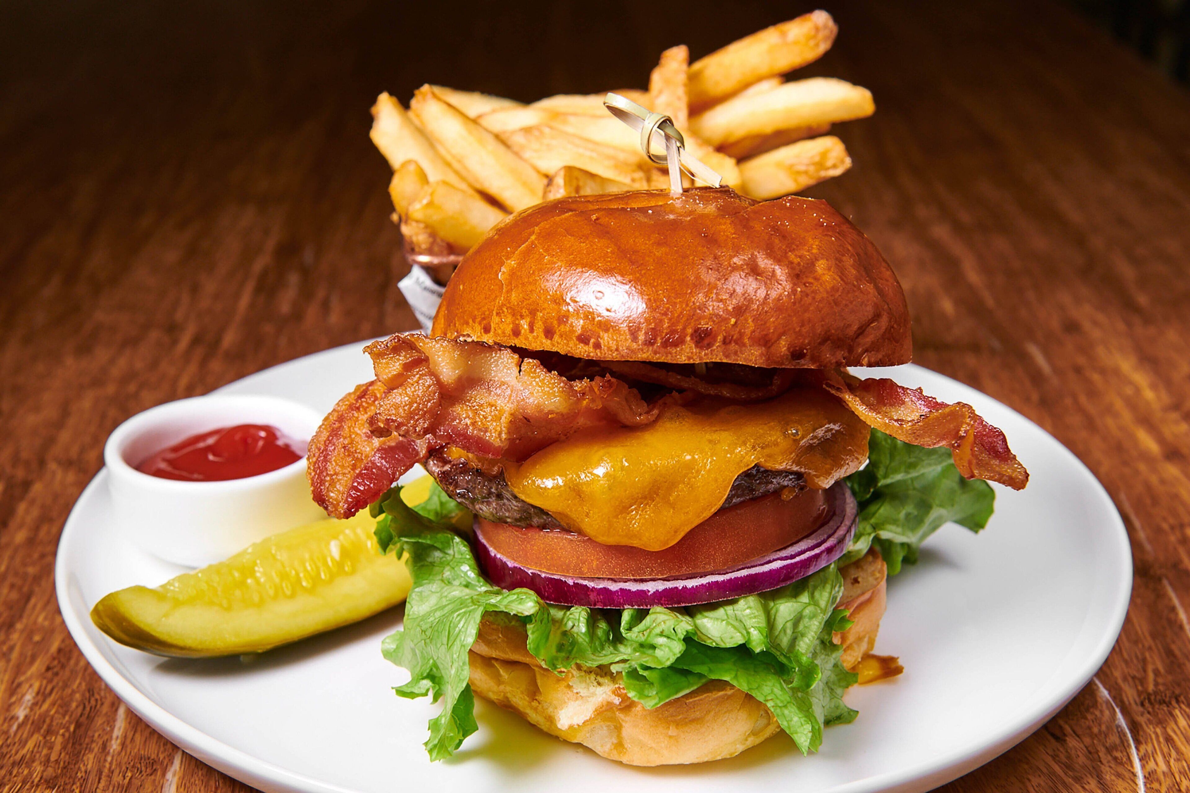 Una hamburguesa con tocino, queso, lechuga y tomate sobre un pan, acompañada de papas fritas y un pepinillo en un plato blanco.