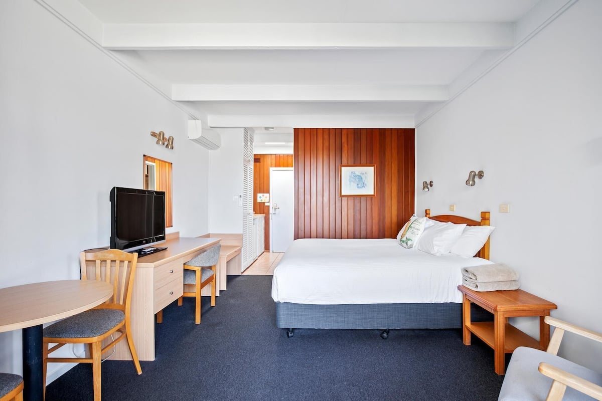A hotel room with a bed, a desk with a TV, a chair, a small table, and a wooden panel wall.