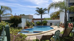 A swimming pool with a curved edge, surrounded by potted plants and palm trees. There are lounge chairs and a small table with a chair. - Banjo Paterson Motor Inn (Lakes Entrance)
