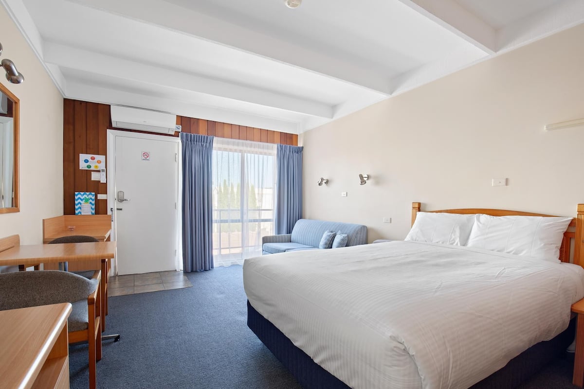 A hotel room with a large bed, a desk, and a chair. There is a door leading to an outdoor area.
