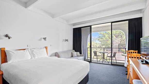 A hotel room with a large bed, a sofa, a desk with chairs, and a balcony with a view of trees and a building. - Banjo Paterson Motor Inn (Lakes Entrance)