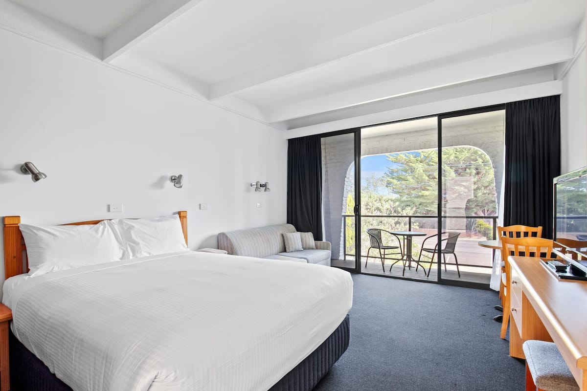 A hotel room with a large bed, a sofa, a desk with chairs, and a balcony with a view of trees and a building.
