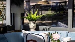 A modern living room with a blue sofa, a decorative plant in a vase, and a view of the street through large windows. - Hôtel Nude Paris - Color Vision (Paris)