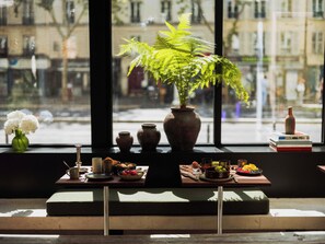 A window with a view of a street, a plant, and a table with food and drink items. - Hôtel Lulu Paris (Paris)
