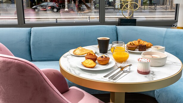 A breakfast setup on a round table with a view of the street through a window. - Hôtel Nude Paris - Color Vision (Paris)