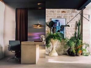 A modern interior with a concrete reception desk, a black office chair, a hanging pendant light, and a variety of potted plants. - Hôtel Lulu Paris (Paris)