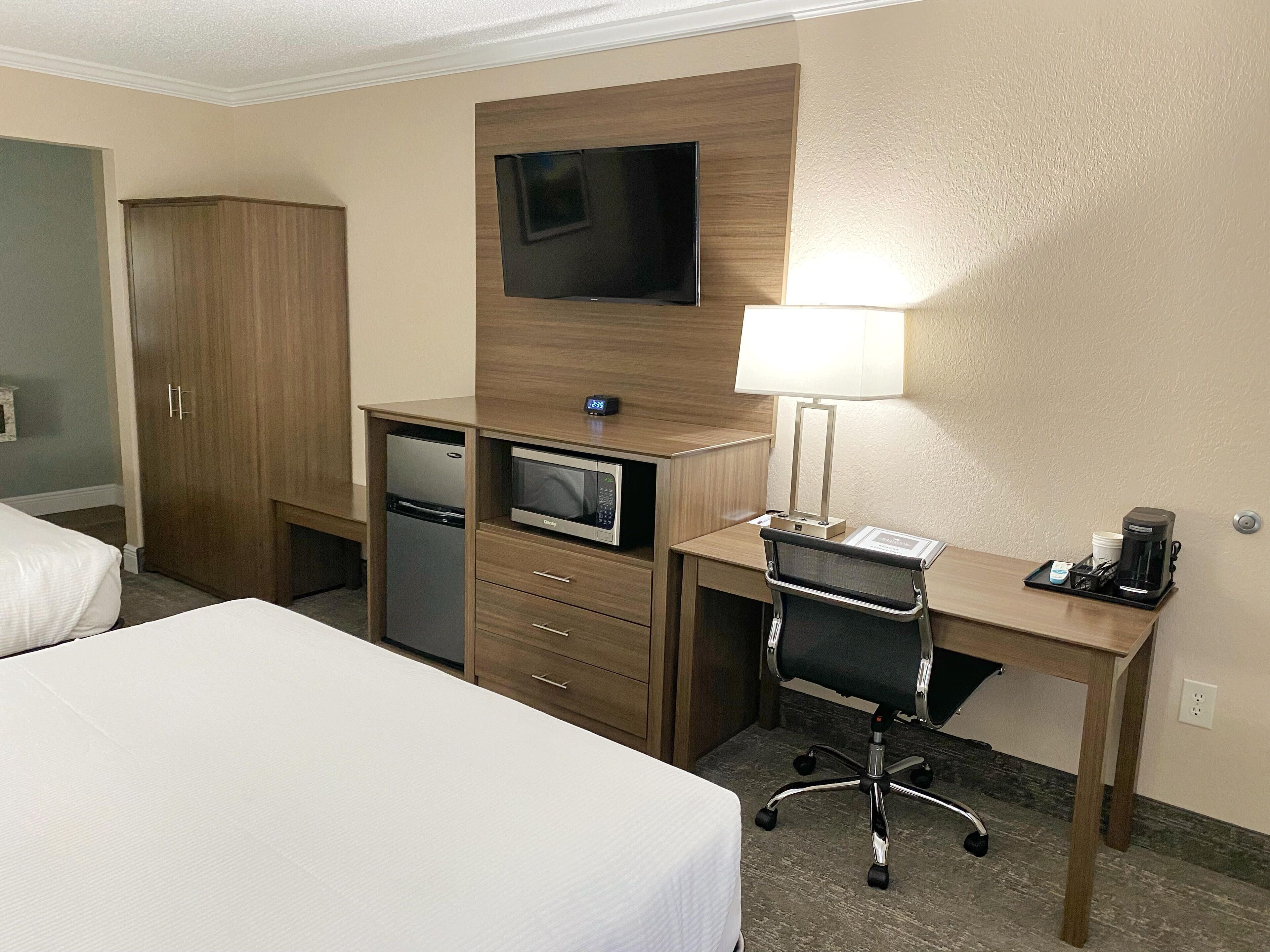 A hotel room with a bed, a desk with a lamp, a microwave, a refrigerator, and a TV on the wall.