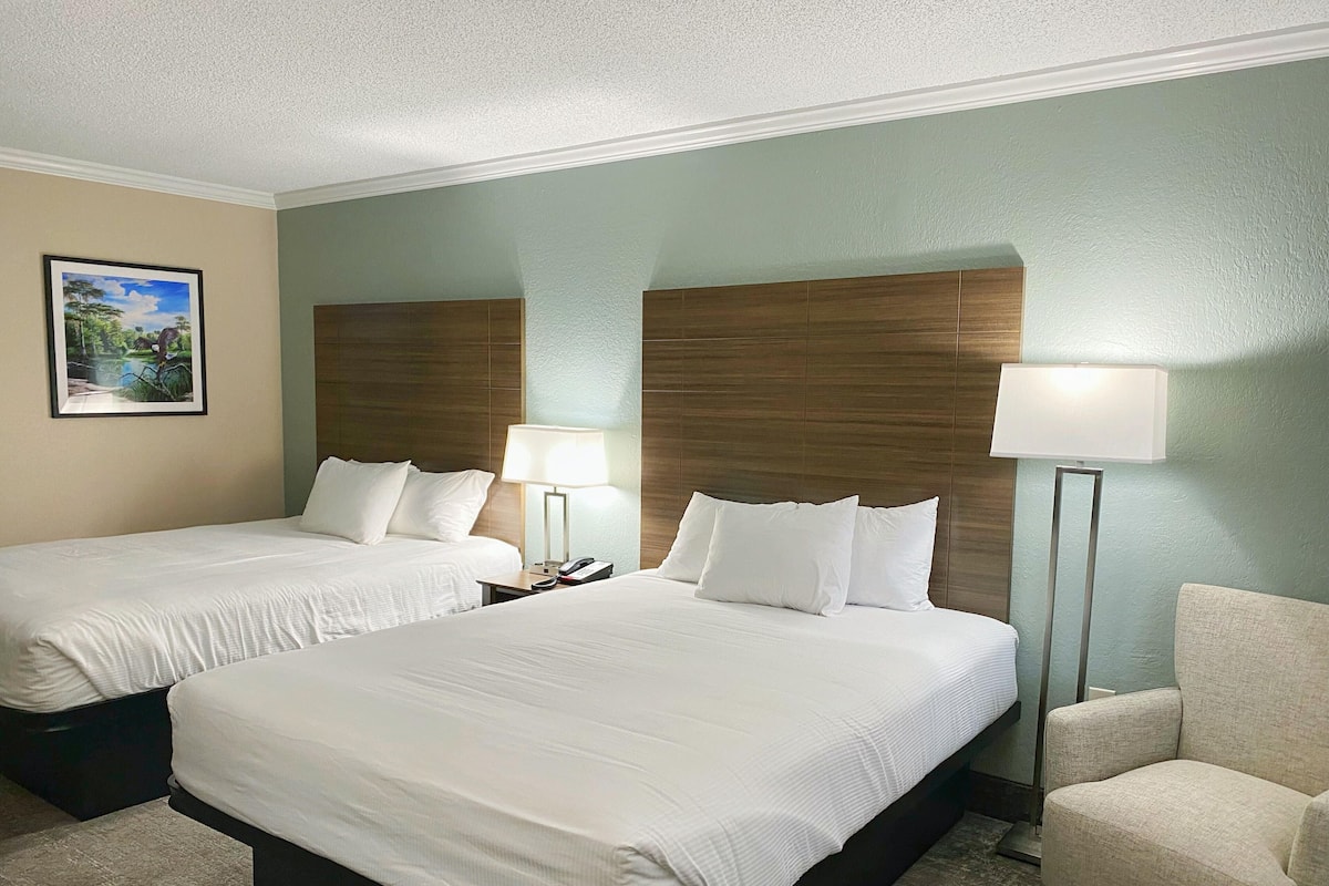 Habitación de hotel con dos camas, una mesita de noche con lámpara, una silla e imagen enmarcada en la pared.