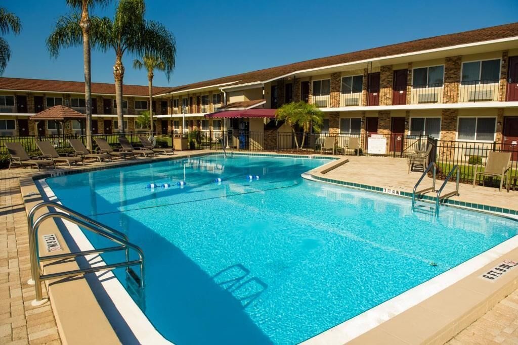 Piscina com espreguiçadeiras e palmeiras, cercada por um edifício de dois andares com varandas.