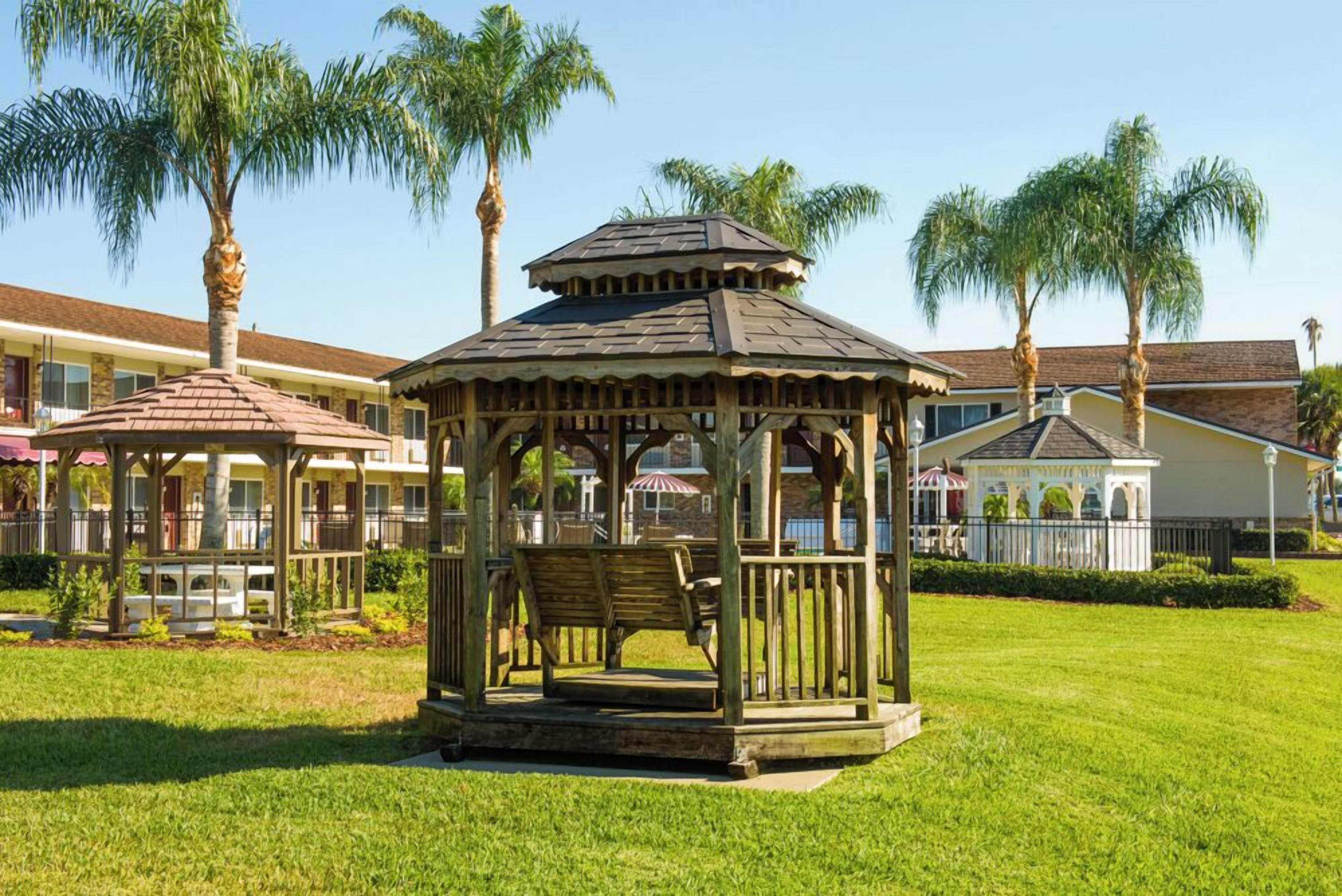 A gazebo with a wooden structure and benches, surrounded by a well-maintained lawn and palm trees, with a building in the background.