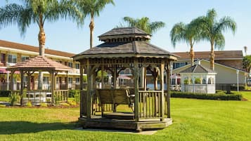 A gazebo with a wooden structure and benches, surrounded by a well-maintained lawn and palm trees, with a building in the background.