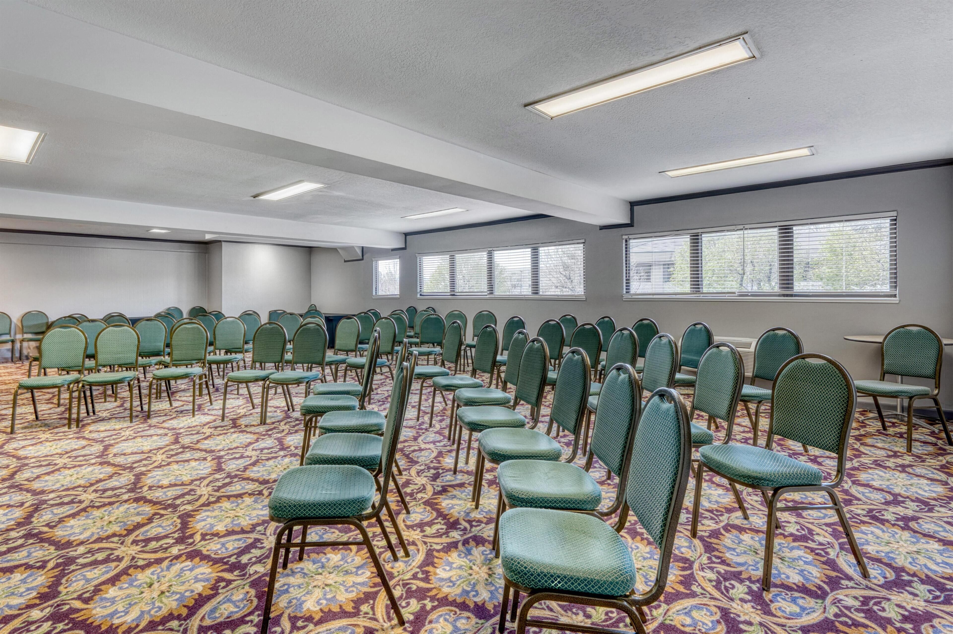 A conference room with rows of green chairs, patterned carpet, and large windows.