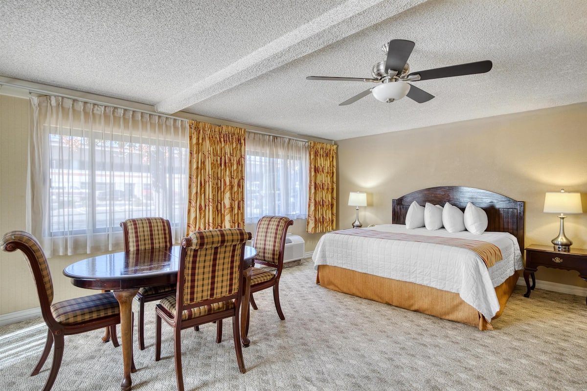 A hotel room with a bed, a dining table with chairs, a ceiling fan, and a window with curtains.