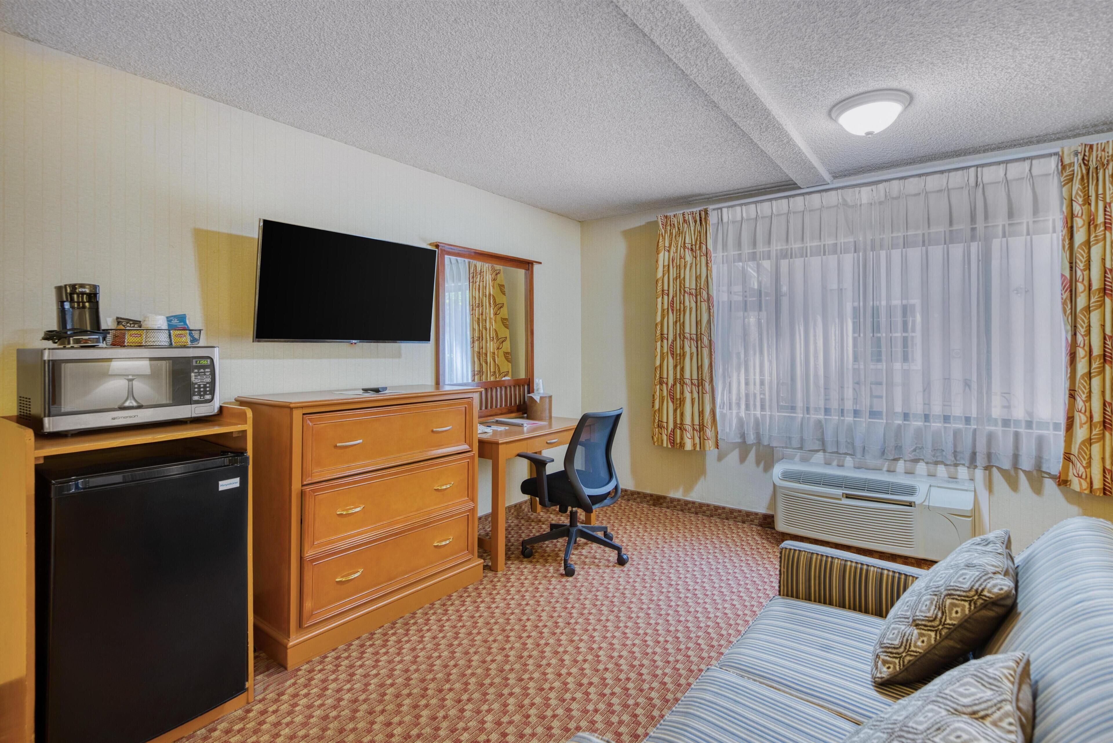 Hotellihuone, jossa on pieni keittiö, työpöytä tuolilla, televisio, ikkuna verhoilla ja sohva.