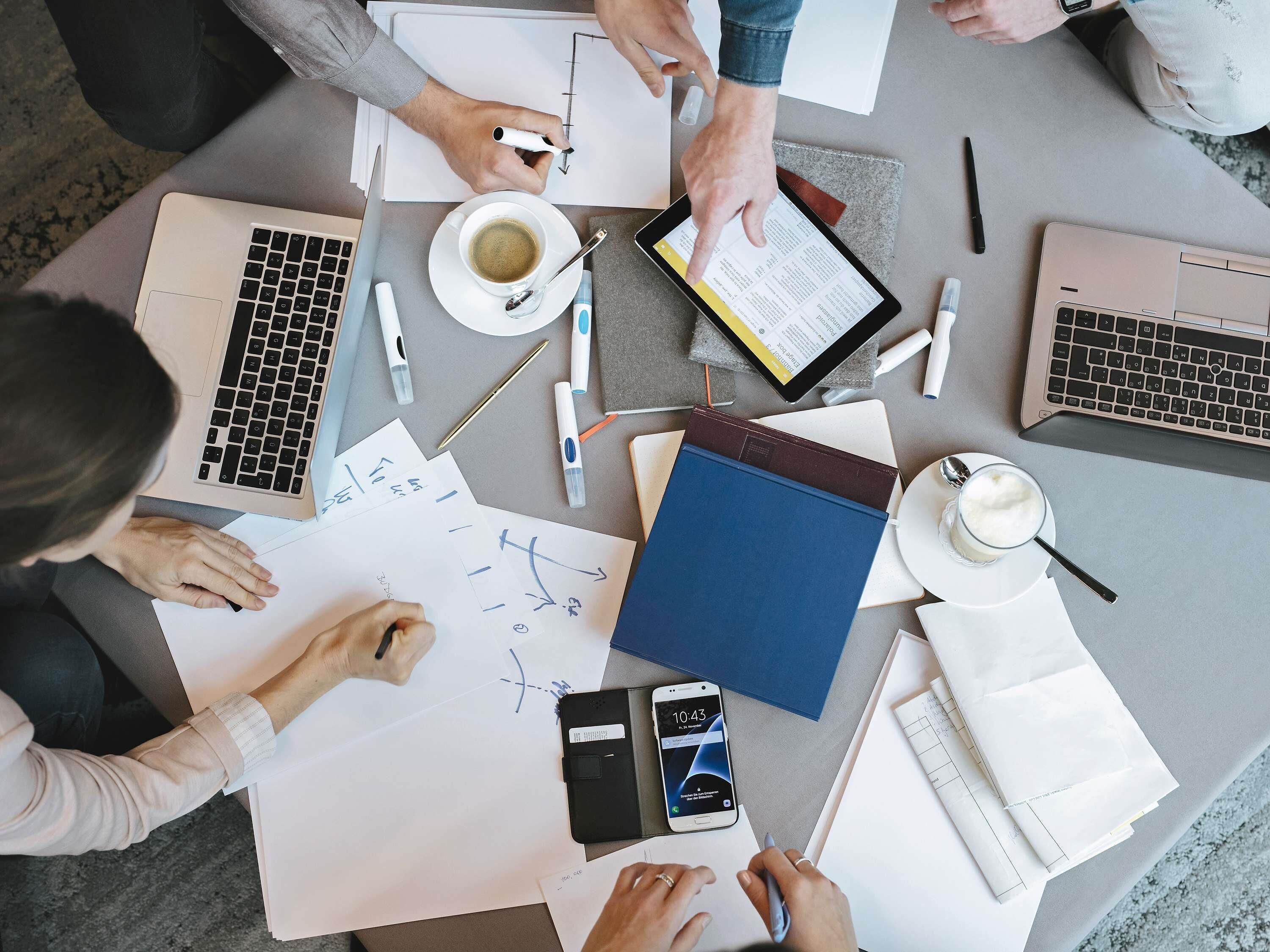 Un grupo de personas en una mesa con laptops, cuadernos y utensilios de escritura, indicando un ambiente de trabajo colaborativo.