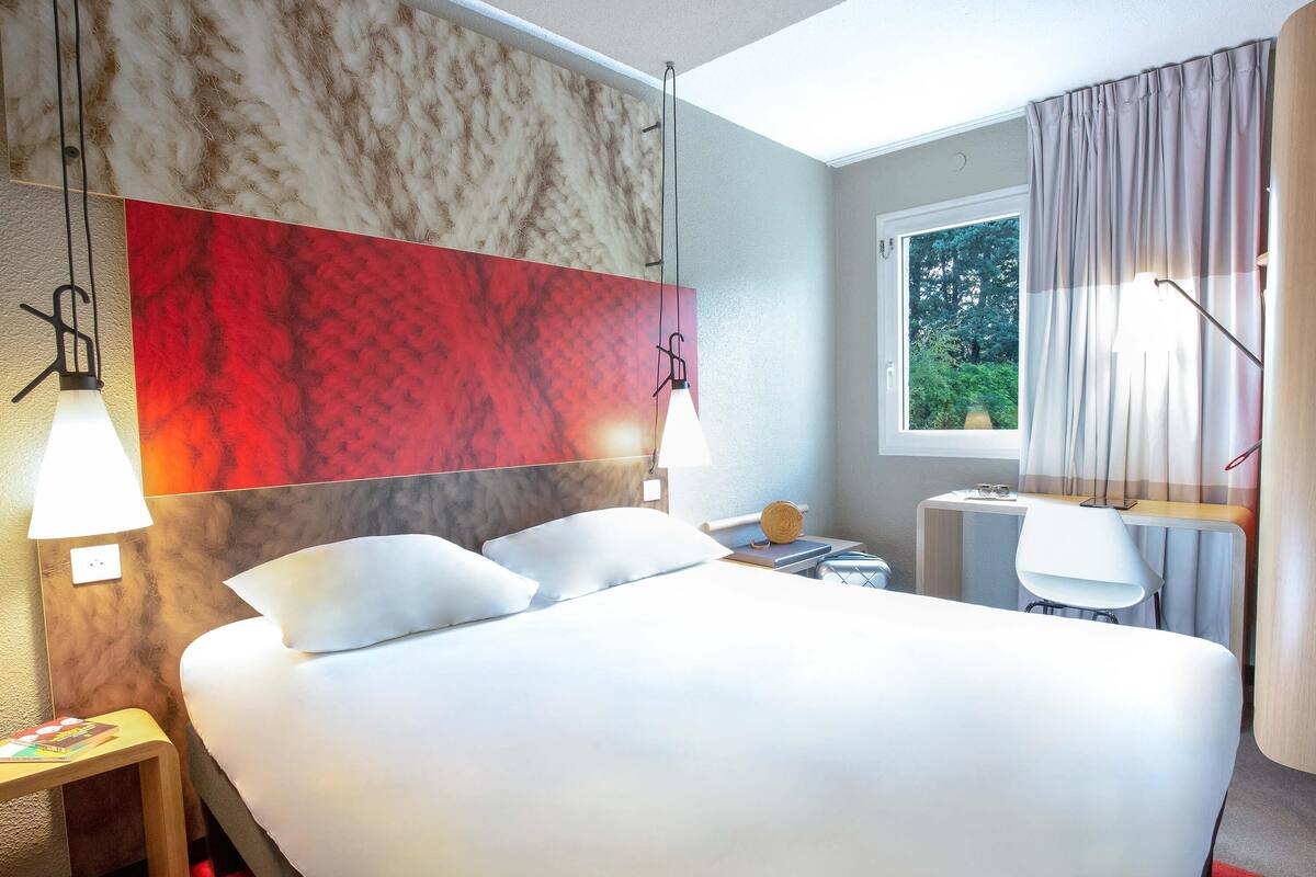 A modern hotel room with a large bed, two bedside lamps, a window with curtains, and a view of greenery outside.