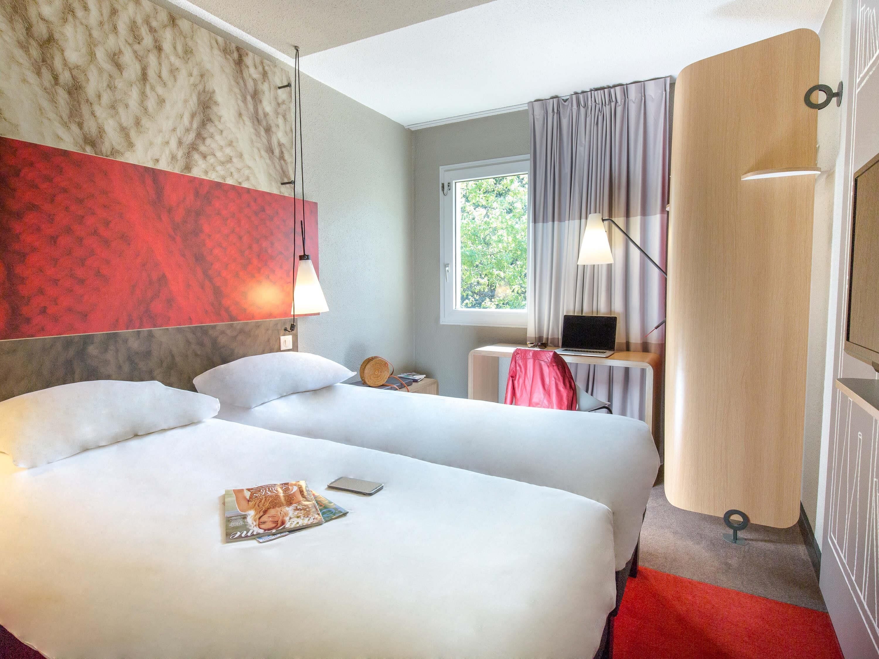 A hotel room with two beds, a desk with a laptop, a window with curtains, and a red carpet.