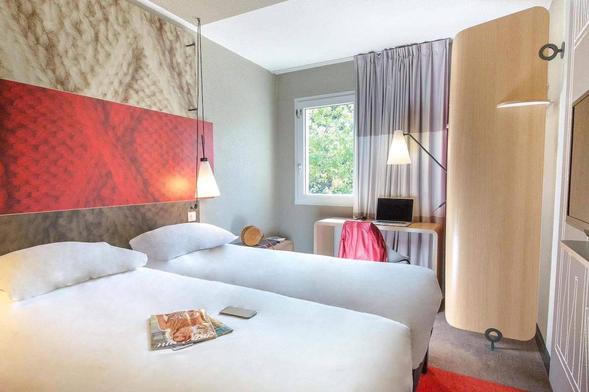 A hotel room with two beds, a desk with a laptop, a window with curtains, and a red carpet.