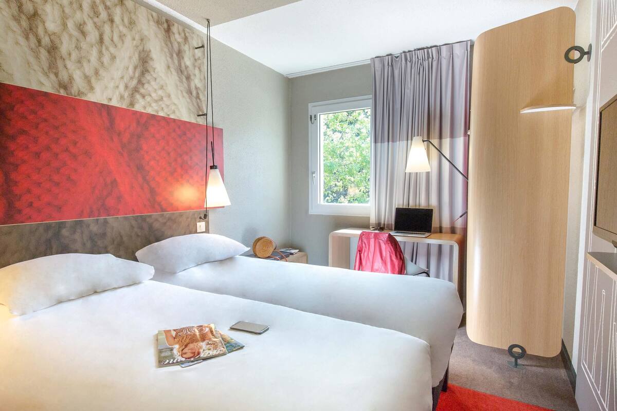 A hotel room with two beds, a desk with a laptop, a window with curtains, and a red carpet.