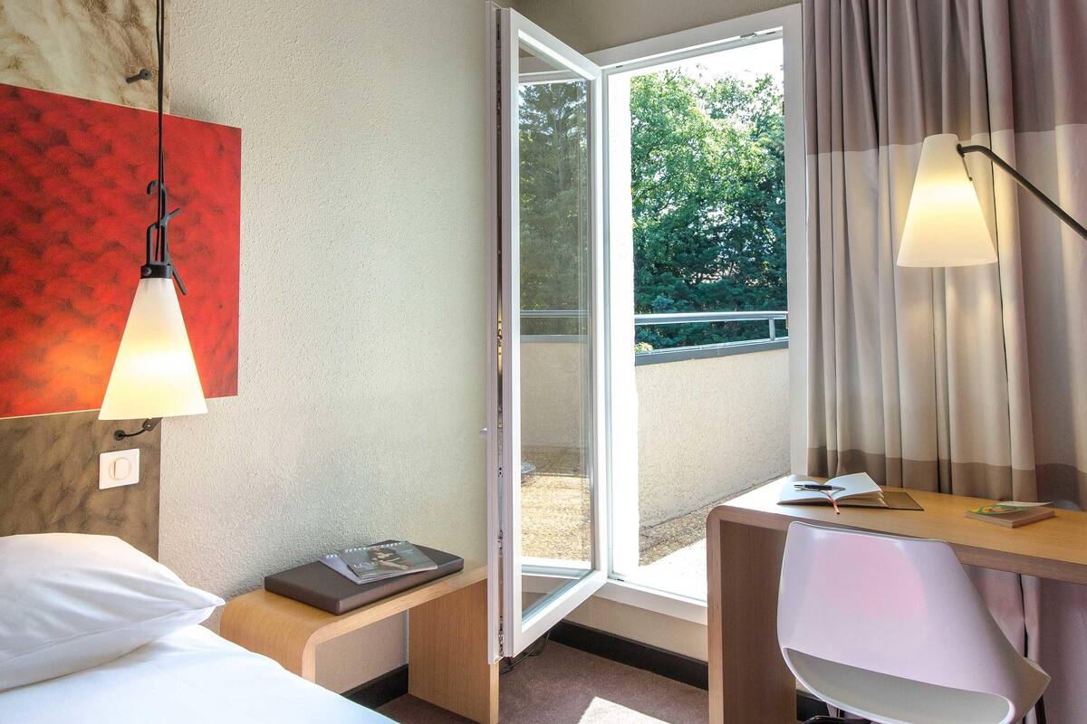 A hotel room with a bed, desk, chair, and a large window with an open door leading to an outdoor area.