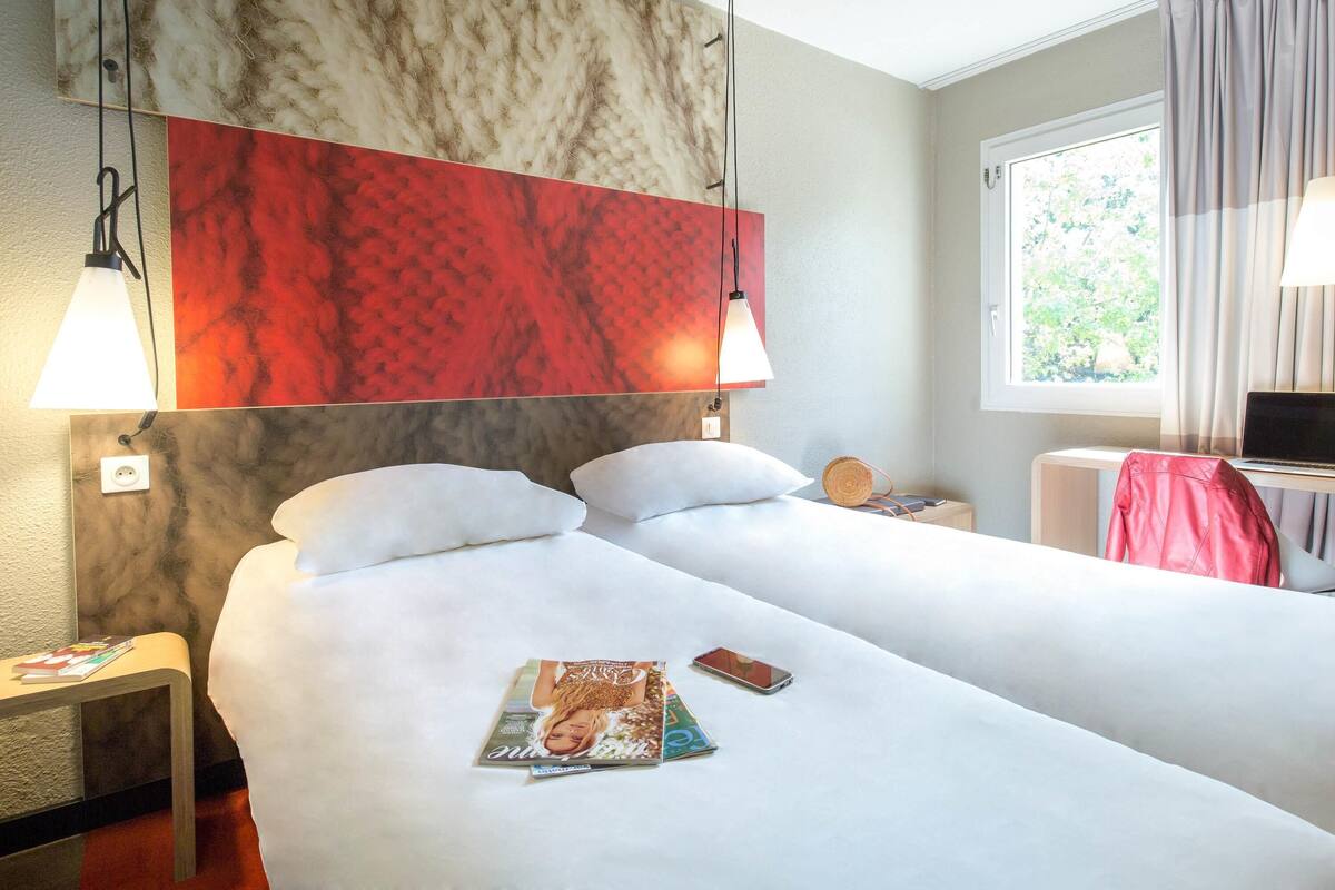 A hotel room with a bed, bedside lamps, a magazine, a smartphone, and a window with curtains.